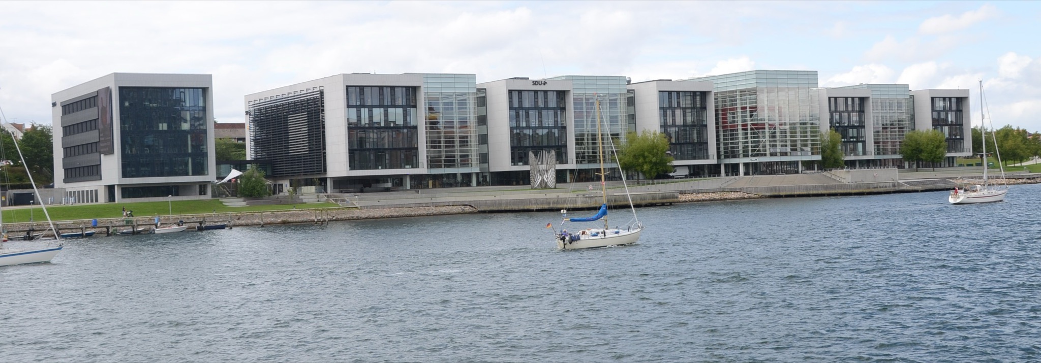 Alsion, the university building in S&oslash;nderborg, seen across the water of Alssund, with a sailing boat in front.