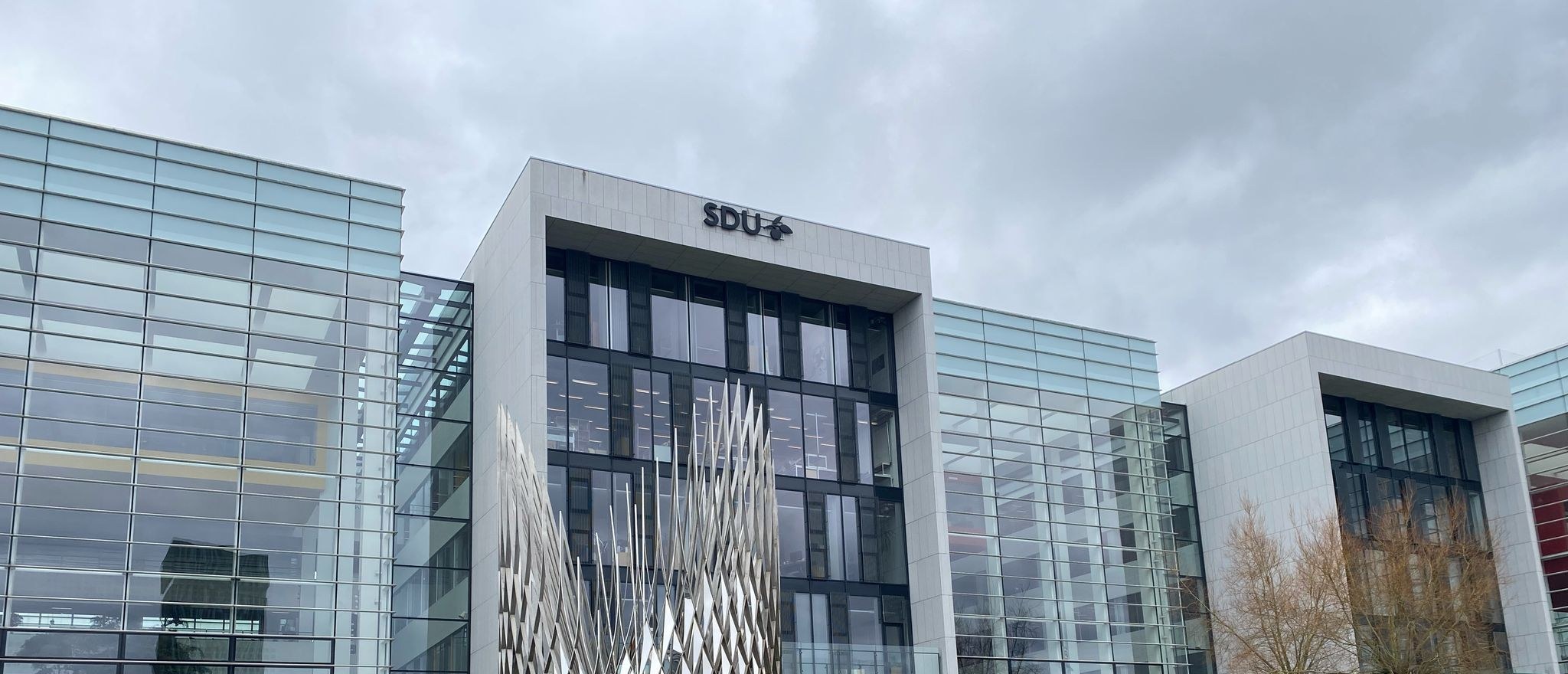 A University of Southern Denmark building in glass and concrete under an overcast sky, with a steel sculpture in front of it.